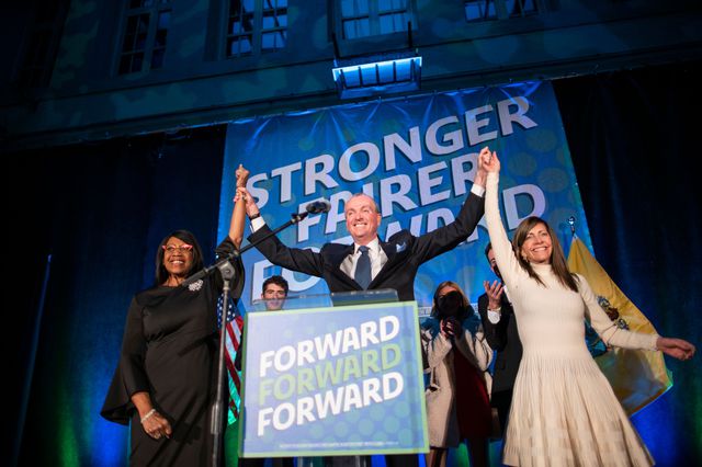 Phil Murphy smiles and raises his arms with his wife and lieutenant cover on the stage, with his "STronger Fairer Forward" slogan behind him